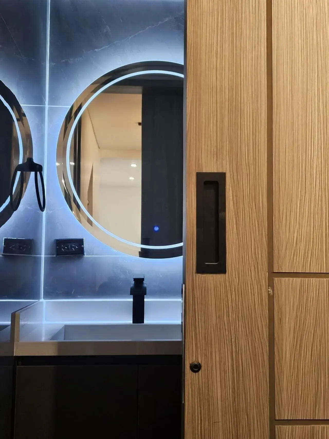 Espejo redondo con luz LED y puerta de madera en un baño