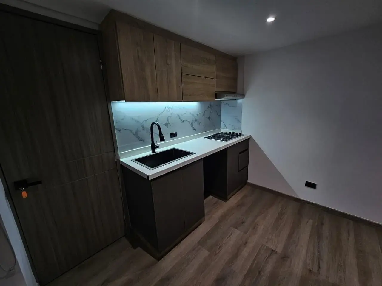 Cocina compacta con mesón negro, salpicadero claro y piso de madera