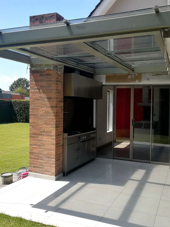 Cubierta de vidrio con estructura metálica sobre una zona social exterior con parrilla, columna de ladrillo y jardín