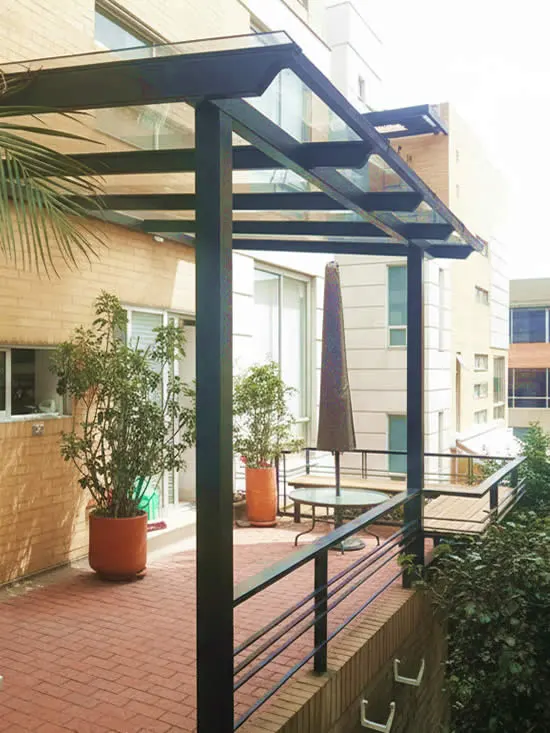 Estructura metálica negra con cubierta de vidrio sobre una terraza con plantas y baranda de metal