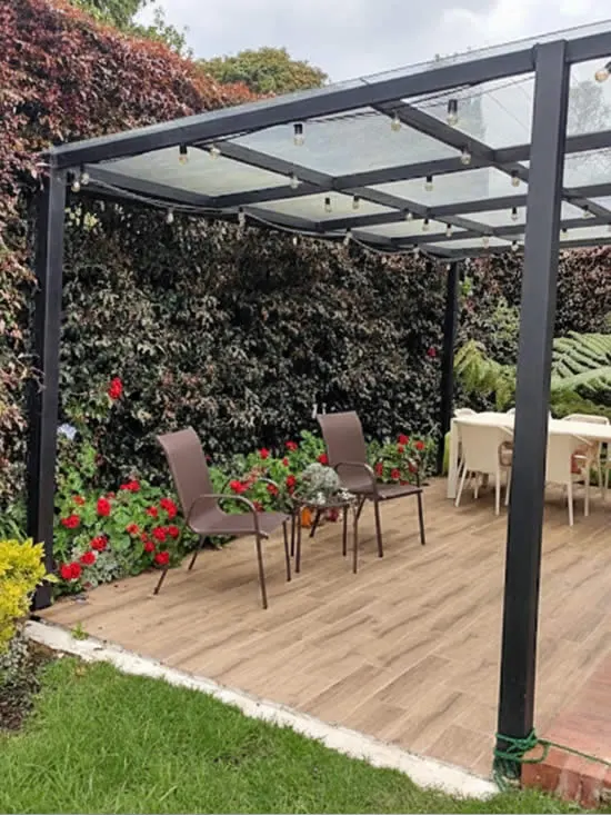 Pérgola de estructura metálica negra con cubierta translúcida sobre una terraza de deck, con sillas y un muro verde al fondo