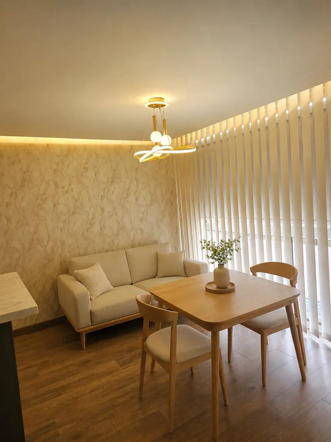 Sala comedor con sofá claro, mesa de madera y cortinas verticales con luz cálida